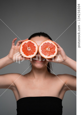 Cheerful woman holding two grapefruit slices over eyes, representing citrus freshness, fun energy and natural beauty 134238304