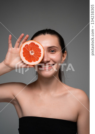 Smiling woman holding grapefruit slice to face, promoting citrus skincare, natural freshness and healthy beauty 134238305