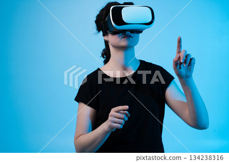 Woman wearing VR headset interacting with virtual environment and pointing forward on blue background Woman wearing VR headset interacting with virtual environment and pointing forward on blue background 134238316
