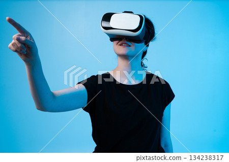 Woman using VR headset and pointing forward on blue background while interacting with virtual interface 134238317