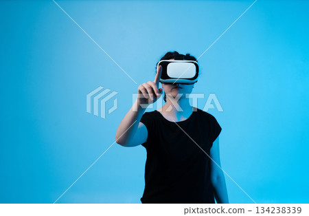 Woman using VR headset and interacting with virtual interface by pointing forward on blue background Woman using VR headset and interacting with virtual interface by pointing forward on blue background 134238339