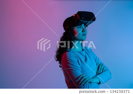 Woman with raised VR headset looking upward thoughtfully in blue pink neon lighting immersive studio scene 134238371