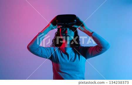 Woman lifting VR headset while transitioning out of virtual environment in dramatic neon blue red lighting 134238373