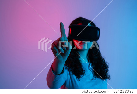 A woman wearing a VR headset reaches forward, interacting with a virtual interface under neon blue and pink lighting. A woman wearing a VR headset reaches forward, interacting with a virtual interface under neon blue and pink lighting. 134238383