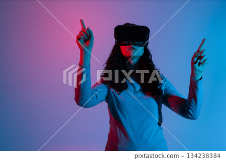 Woman with VR headset raising hands interacting with immersive digital environment under neon lights Woman with VR headset raising hands interacting with immersive digital environment under neon lights 134238384