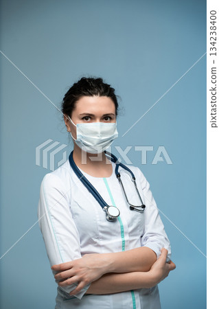 Female medical professional in mask and white uniform with stethoscope posing on blue background Female medical professional in mask and white uniform with stethoscope posing on blue background 134238400