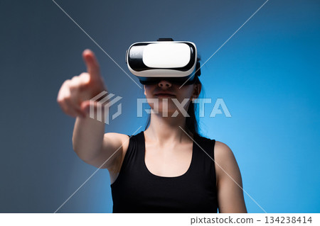 Young woman reaching forward while using VR headset in immersive digital environment with blue lighting Young woman reaching forward while using VR headset in immersive digital environment with blue lighting 134238414