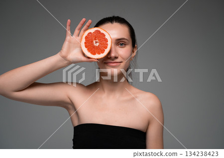 Positive young woman holding a grapefruit slice over her eye, highlighting citrus freshness, natural skincare Positive young woman holding a grapefruit slice over her eye, highlighting citrus freshness, natural skincare 134238423