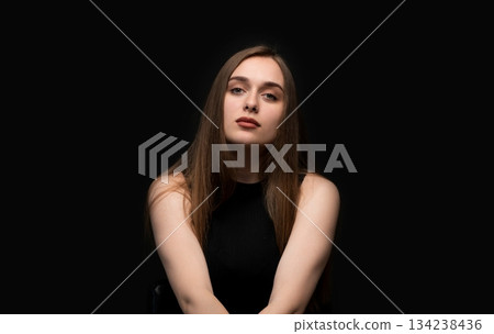 Calm studio portrait of a young woman with neutral facial expression against a dark background Calm studio portrait of a young woman with neutral facial expression against a dark background 134238436