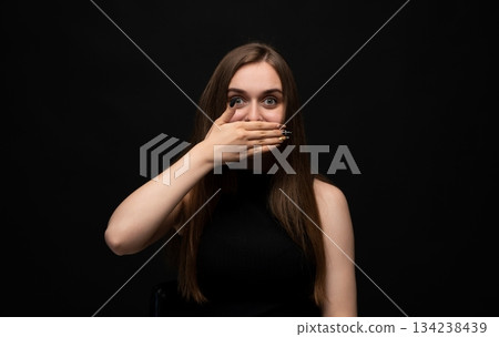 Surprised young woman covering mouth with hand in shocked emotional reaction on dark studio background 134238439