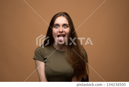 Funny young woman sticking out tongue with wide playful eyes in expressive studio portrait on brown background 134238505