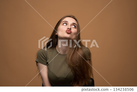 Playful young woman puckering lips upward in cute kissing gesture on warm brown studio portrait background 134238506