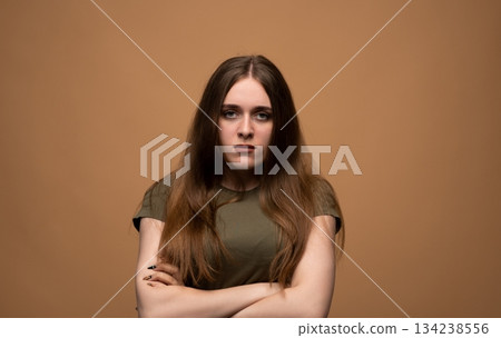 Serious young woman with crossed arms showing confident strict expression in warm studio portrait setting Serious young woman with crossed arms showing confident strict expression in warm studio portrait setting 134238556