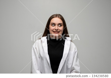 Young woman making nervous grimace with tense smile in studio portrait on gray background 134238580