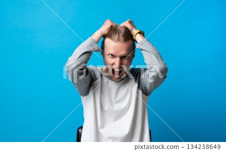 Angry young man pulling his hair in frustration on blue background, emotional stress and rage expression Angry young man pulling his hair in frustration on blue background, emotional stress and rage expression 134238649