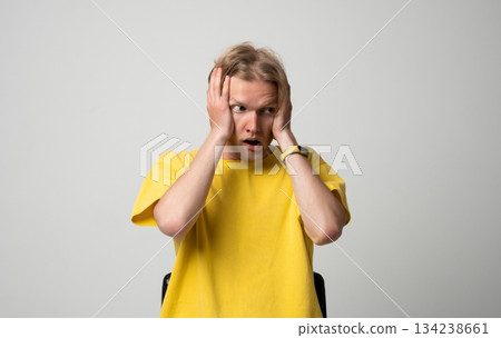 Shocked young man holding his cheeks with both hands and reacting with wide eyes in neutral studio setting Shocked young man holding his cheeks with both hands and reacting with wide eyes in neutral studio setting 134238661