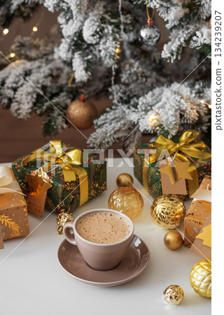 Cup of Hot Drink with Christmas Gifts and Ornaments in Cozy Holiday Setting against Christmas Tree and Lights Cup of Hot Drink with Christmas Gifts and Ornaments in Cozy Holiday Setting against Christmas Tree and Lights 134239207