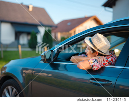 Traveler woman in red ethnic dress and hat drives car through scenic countryside of Croatia in summer. Surrounded by lush fields and forests, she enjoys peaceful and adventurous road trip 134239343