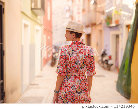Woman in dress strolls through colorful streets of Spanish coastal town of La Vila Joiosa . sunny winter atmosphere highlights charm of Mediterranean architecture and quiet seaside life, back view Woman in dress strolls through colorful streets of Spanish coastal town of La Vila Joiosa . sunny winter atmosphere highlights charm of Mediterranean architecture and quiet seaside life, back view 134239346