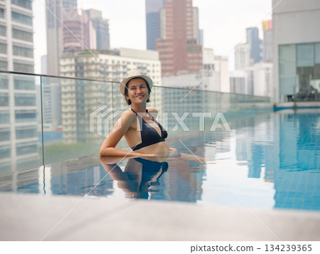 Young asian woman relaxing by pool at Kuala Lumpur hotel with view of surrounding skyscrapers, enjoying leisure time in vibrant urban setting. Young asian woman relaxing by pool at Kuala Lumpur hotel with view of surrounding skyscrapers, enjoying leisure time in vibrant urban setting. 134239365