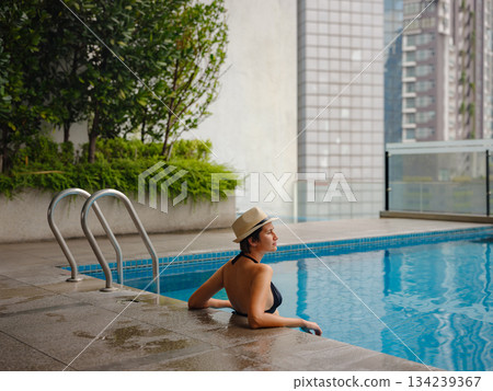 Young asian woman relaxing by pool at Kuala Lumpur hotel with view of surrounding skyscrapers, enjoying leisure time in vibrant urban setting. 134239367