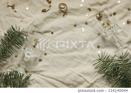 Golden star confetti tinsel, beige linen table cloth background. Champagne glasses. Fir branches floral frame, copy space. Christmas party concept. New Years flatlay, top view. Festive holiday banner Golden star confetti tinsel, beige linen table cloth background. Champagne glasses. Fir branches floral frame, copy space. Christmas party concept. New Years flatlay, top view. Festive holiday banner 134240044