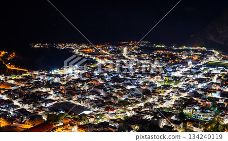 City lights of Banos de Agua Santa shine in the Andean valley at night in Ecuador. Panoramic urban landscape seen from Mirador de la Virgen features illuminated streets and dark mountains City lights of Banos de Agua Santa shine in the Andean valley at night in Ecuador. Panoramic urban landscape seen from Mirador de la Virgen features illuminated streets and dark mountains 134240119