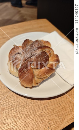 Freshly baked cinnamon bun served on a white plate on a wooden table in a cozy cafe setting. The image represents comfort food, homemade baking, warmth and simple everyday pleasure Freshly baked cinnamon bun served on a white plate on a wooden table in a cozy cafe setting. The image represents comfort food, homemade baking, warmth and simple everyday pleasure 134240597