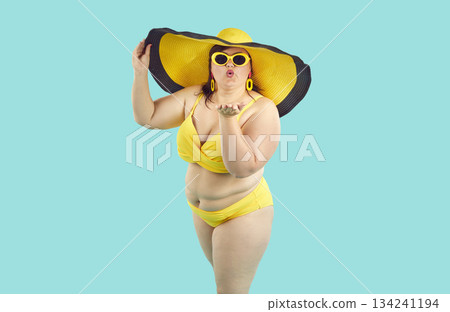 Fat woman in bikini, sunglasses and big beach hat giving air kiss on studio blue background. 134241194