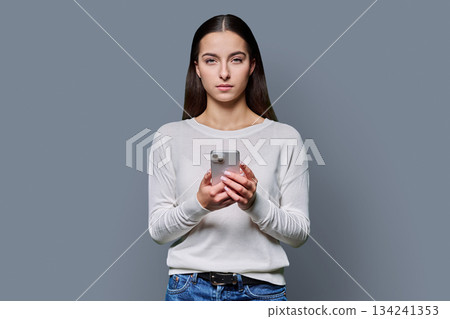 Portrait of serious teenage girl holding smartphone on gray studio background 134241353