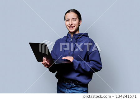 Portrait of smiling teenage girl holding laptop on gray studio background 134241571