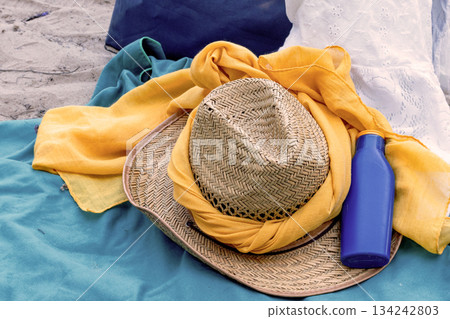 Beach day accessories lying on a blanket for sun protection and relaxation, featuring a straw hat, a bright yellow scarf, and a blue bottle of sunscreen Beach day accessories lying on a blanket for sun protection and relaxation, featuring a straw hat, a bright yellow scarf, and a blue bottle of sunscreen 134242803