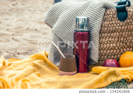 Relaxing outdoor picnic setup on the sand, featuring a thermos, mate gourd with bombilla, woven bag, and fresh fruits, enjoying leisure time 134242808