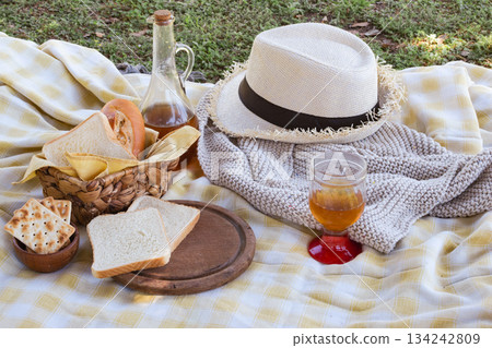 Checkered picnic blanket on green grass with straw hat, bottle and glass of amber drink, crackers and assorted breads in a basket for a relaxing summer outdoor snack Checkered picnic blanket on green grass with straw hat, bottle and glass of amber drink, crackers and assorted breads in a basket for a relaxing summer outdoor snack 134242809