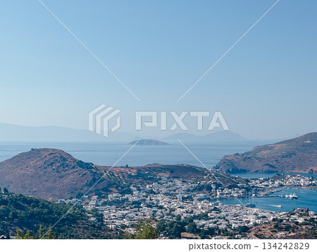 Greek island town of whitewashed buildings cascading to a busy harbor, boats bobbing in turquoise aegean waters beneath sunlit hills and distant mountains perfect travel escape 134242829