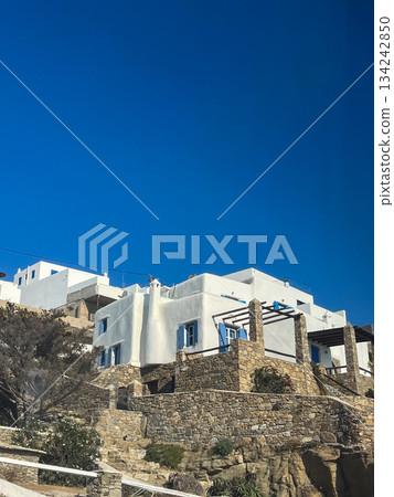 Mykonos island architecture featuring traditional cycladic whitewashed buildings with blue shutters and stone walls climbing a rugged hillside under a vibrant clear sky Mykonos island architecture featuring traditional cycladic whitewashed buildings with blue shutters and stone walls climbing a rugged hillside under a vibrant clear sky 134242850
