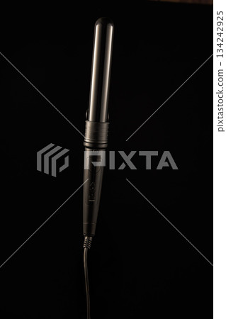 A hair curling iron is shown with a focus on its sleek design. The curling iron is turned off and positioned against a dark surface ready for use in creating waves or curls. 134242925