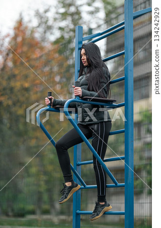 brunette woman is working out at a stadium. She is using a pull up bar while dressed in black workout clothes. Trees are in the background showing the season. 134242929