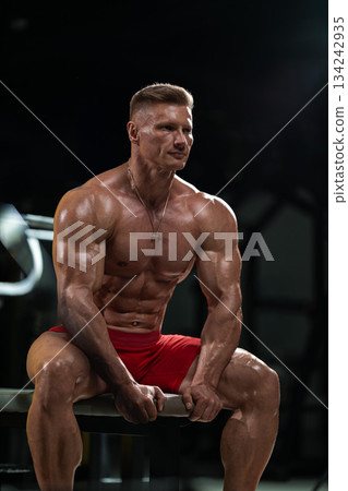 athlete sits on a bench in a gym. He holds weights in his hands while focusing on his workout routine. The lighting highlights his physique during training. 134242935