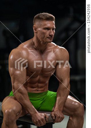 muscular athlete sits on a bench in a gym. He appears to be resting between sets while focusing on his workout. The environment is equipped for strength training. 134242936