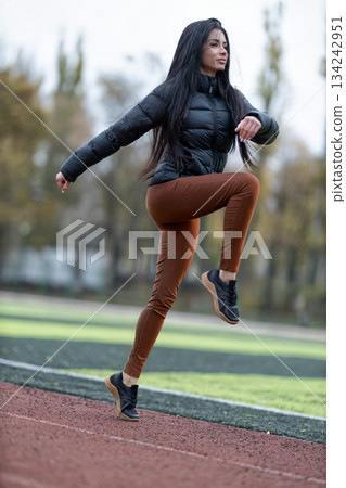 brunette woman runs on the track at a stadium. She wears a black jacket and brown pants. The scene shows her in motion with a focus on her fitness activity. brunette woman runs on the track at a stadium. She wears a black jacket and brown pants. The scene shows her in motion with a focus on her fitness activity. 134242951