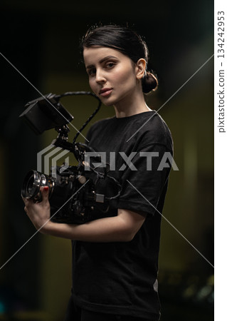 Brunette stands holding a camera in both hands while looking at the viewer. The setting is indoors during the evening with good lighting. Her shirt is black. 134242953