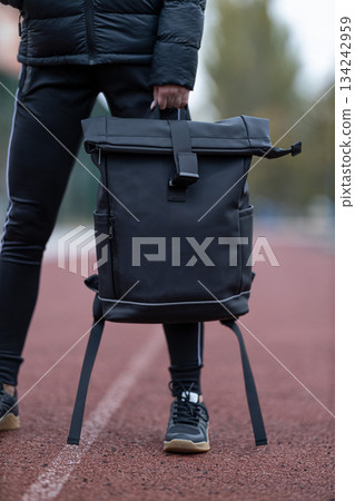 brunette wears a black jacket and holds a black backpack. She stands on a running track during early morning. The surrounding area shows trees and sports equipment. 134242959