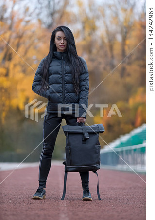 Brunette woman in black jacket and black backpack stands confidently on a track. Trees in the background show colorful leaves. It is a sunny day in fall season. 134242963