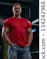 man with a muscular build stands near a Smith machine in a gym. He is wearing a red shirt and has his hands in his pockets. The lighting highlights his features. 134242966