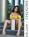brunette woman in a yellow dress is sitting on the steps. She holds a box while enjoying the sunlight. The setting features modern architecture and greenery nearby. 134242968