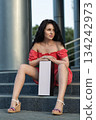 woman with brunette hair sits on dark steps. She wears a red dress and is holding a box. The setting is outdoors in daylight with structures in the background. 134242973