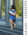 brunette woman in a dress walks down steps while carrying a box. The setting is an outdoor area with modern architecture and greenery. It is daytime. 134242974