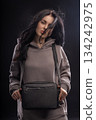 brunette woman is wearing a hoodie and holding a bag while posing in a dimly lit studio. Her hair is flowing around her as she looks at the camera. 134242975