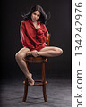 brunette woman wearing a hoodie is sitting on a wooden chair with her legs stretched out. She is posing with her hands creating an artistic look in a studio setting. 134242976